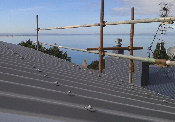 A close-up view of a residential roof featuring TRS 7 steel roofing profile with visible fasteners, overlooking a scenic coastal background.