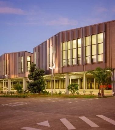 A modern commercial building with large glass windows and dark corten steel cladding, illuminated at dusk.