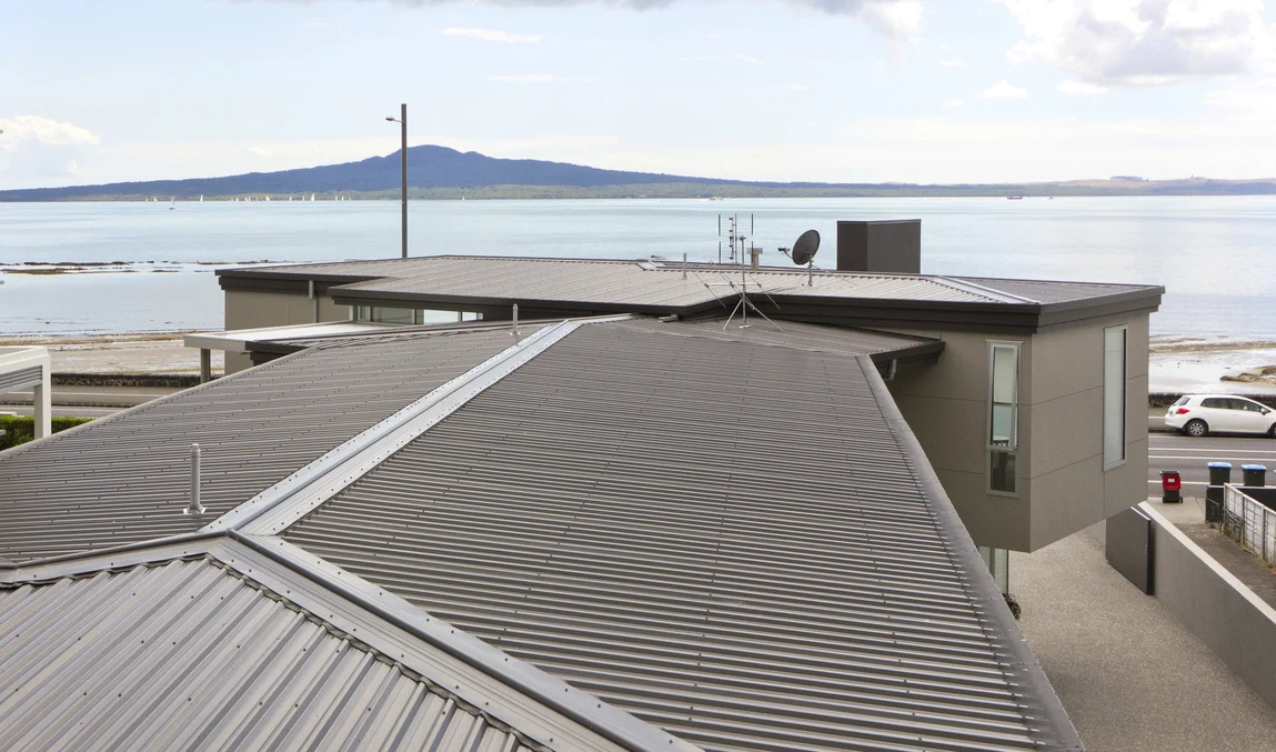 A waterfront home with corrugated metal roofing and a scenic view of the ocean and distant hills.