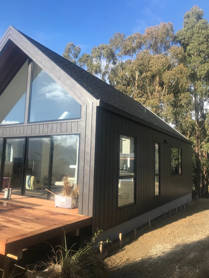 A small modern home with dark steel cladding and large glass windows surrounded by natural greenery.