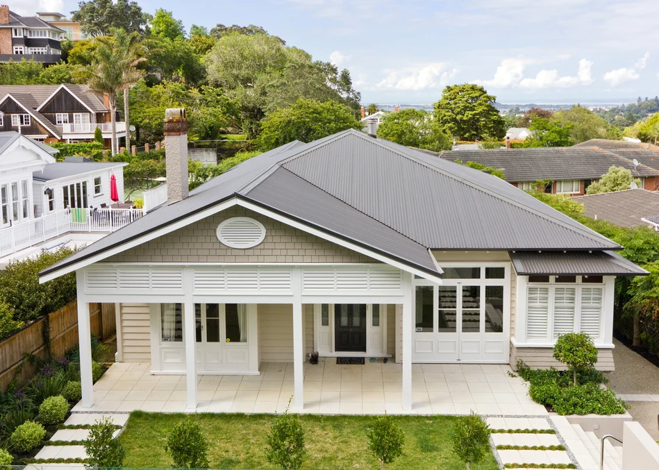 A modern single-storey home with a dark metal roof and light-coloured walls, surrounded by lush greenery.