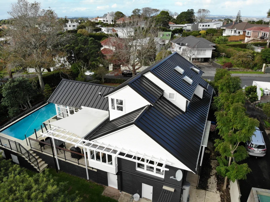 A white house with a black standing seam metal roof, surrounded by lush greenery and a pool.