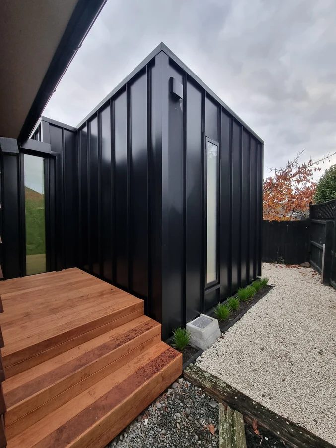 A minimalist home featuring dark vertical cladding, timber steps, and a gravel pathway with greenery.