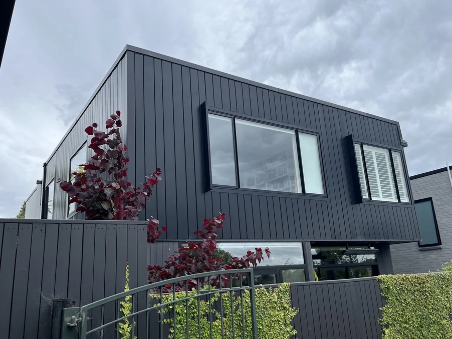 A modern two-storey home with sleek black vertical cladding, surrounded by vibrant plants and a cloudy sky.