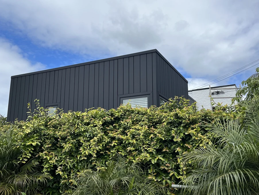 A modern home with dark vertical cladding surrounded by lush green foliage under a cloudy sky.