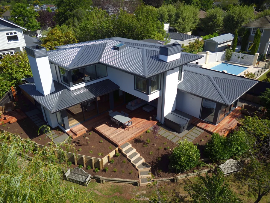 A modern two-storey home with grey metal roofing, expansive wooden decking, and landscaped gardens.