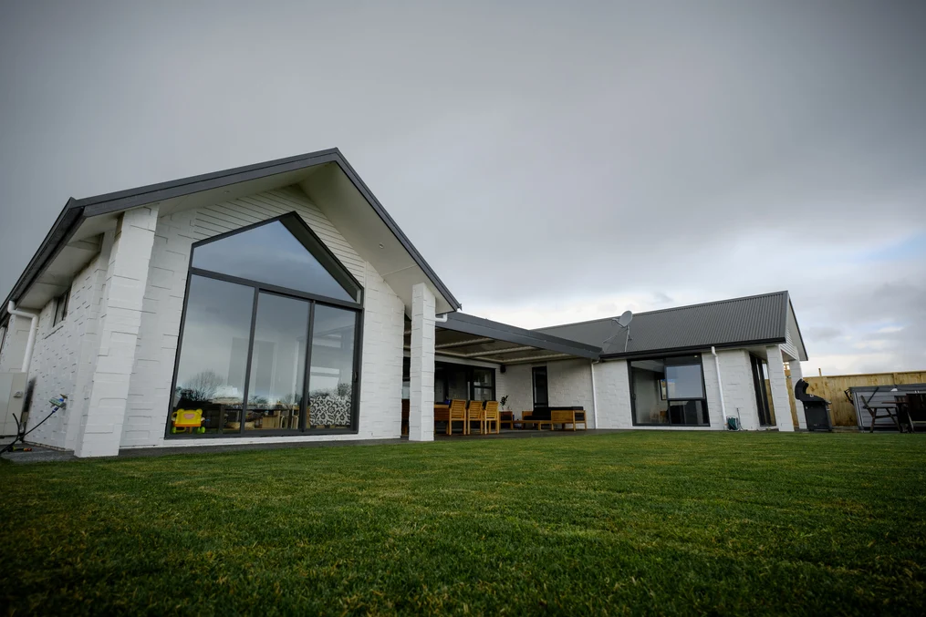 A modern single-storey home with a grey metal roof and large glass windows, surrounded by a lush green lawn under an overcast sky.