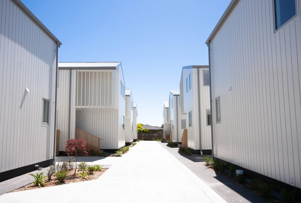 A modern housing complex with light-coloured vertical cladding and landscaped pathways under a bright blue sky.