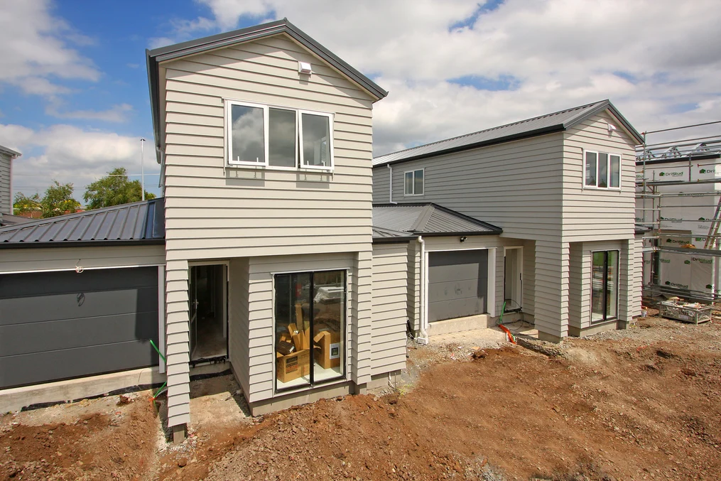 A pair of newly constructed light-coloured townhouses with grey metal roofing, under a partly cloudy sky.