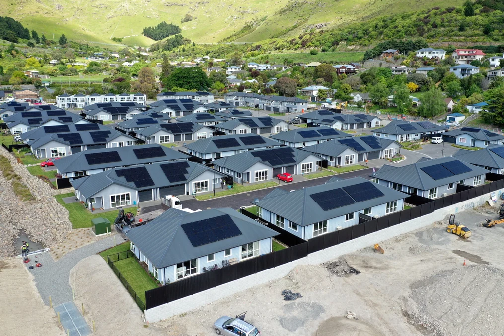 A community of single-storey homes with grey metal roofs and solar panels, set against a lush green hillside.