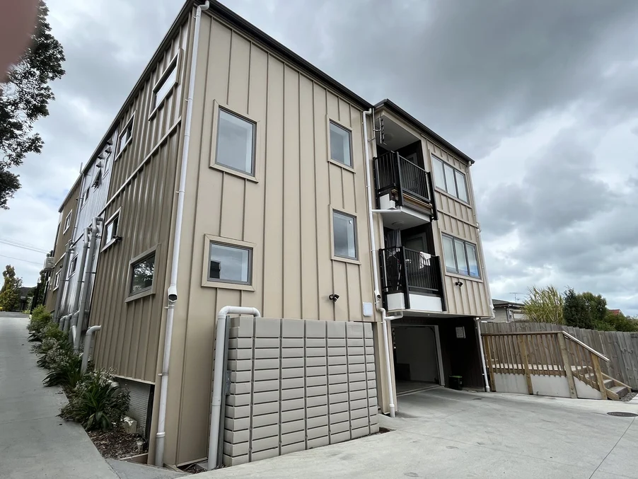 A three-storey residential building with beige vertical cladding and black accents under a cloudy sky.