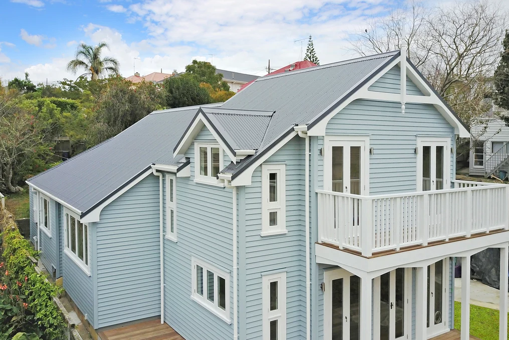 two-storey light blue house with white accents and a grey corrugated metal roof, set in a landscaped suburban neighbourhood.